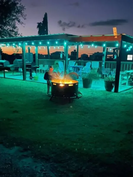 A fire pit glowing on the lawn at dusk in front of the pavilion, lit with teal string lights